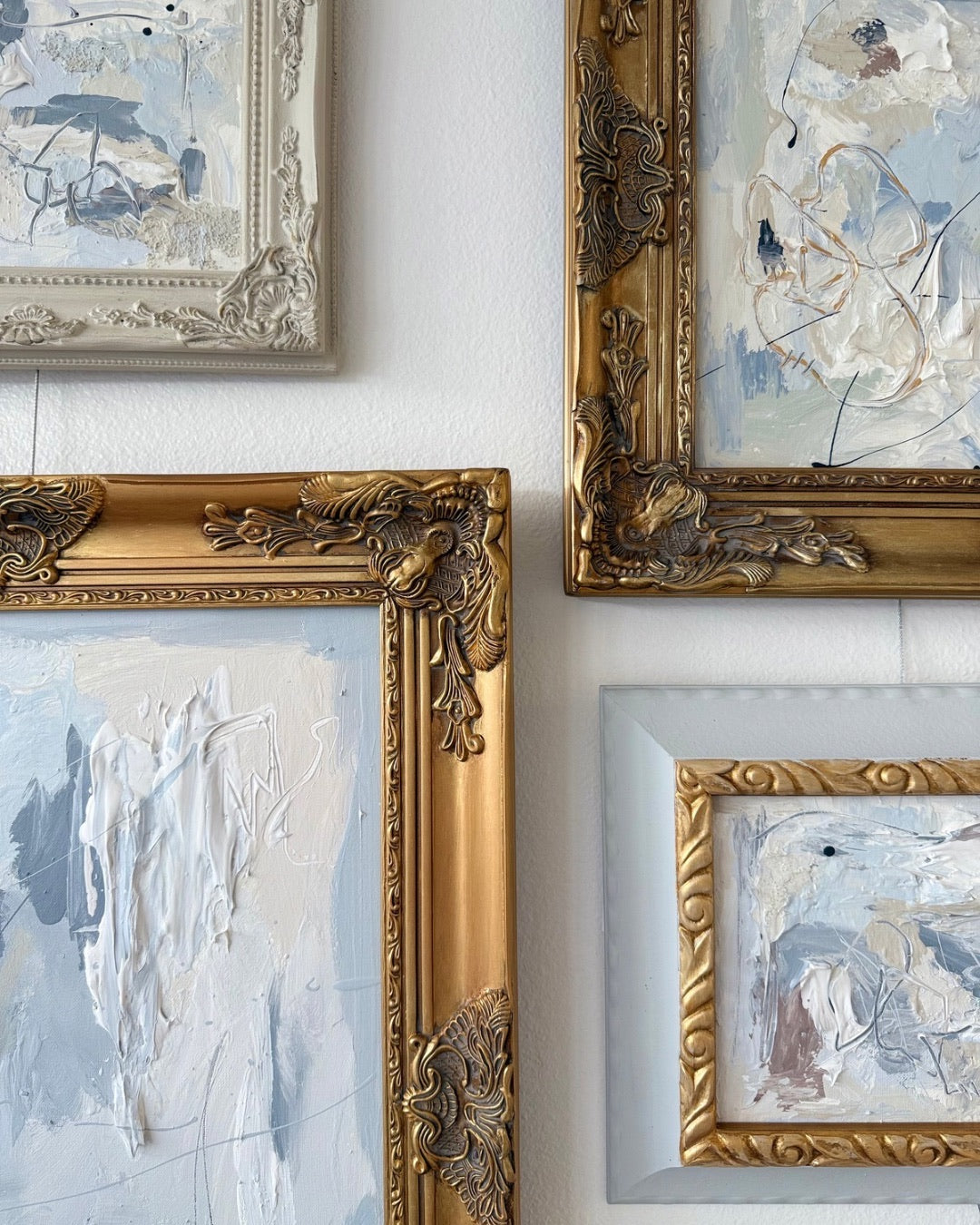 Four pieces of abstract art in ornate gold frames on a white wall.